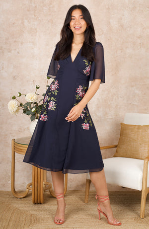 Ready to have heads turning, this classic design features elegant floral embroidery with pops of green, pink and white across the front of its navy blue base. The vintage-inspired silhouette offers a front panel, plus a tie back detail under the bust, perfect for flattering all figures. This dress is complete with delicate, semi-sheer flutter sleeves and a flowing midi length skirt. The perfect choice for those looking for a dream garden party dress this spring. 